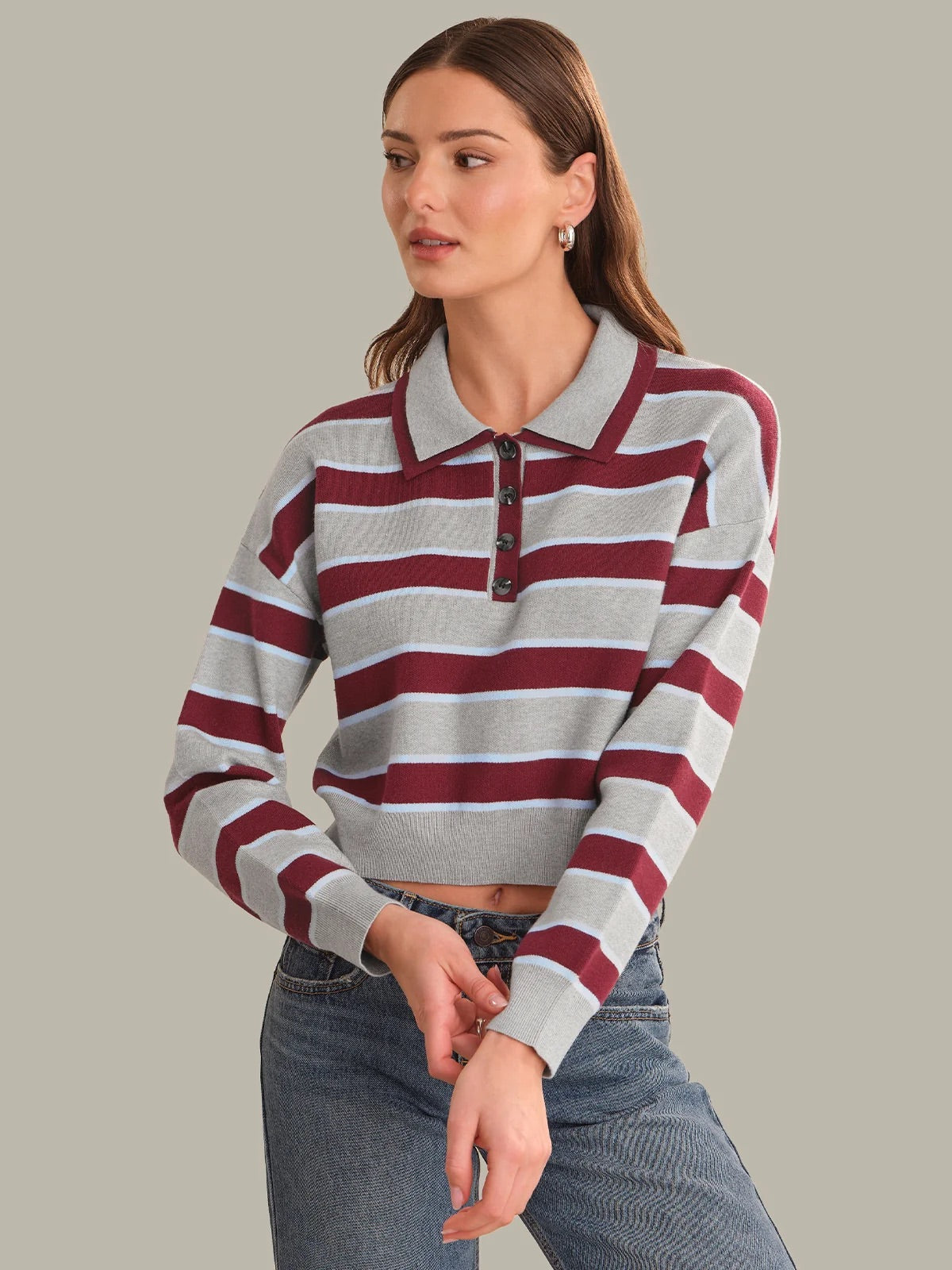 A woman wearing a cropped long-sleeve polo shirt with horizontal gray and burgundy stripes, paired with blue jeans, against a solid light gray background.