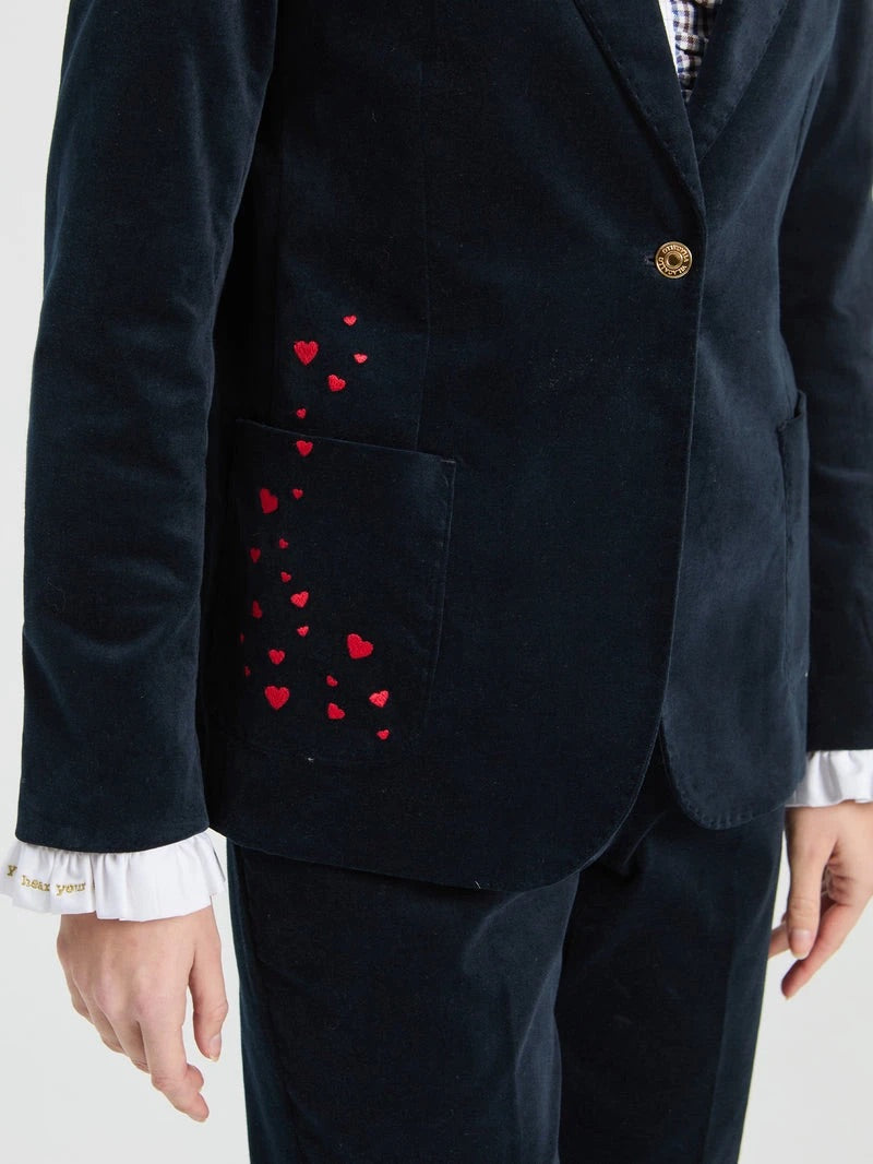 A close-up of a dark navy velvet blazer with red embroidered hearts on the pocket, worn by a model with a white ruffled shirt cuff visible.