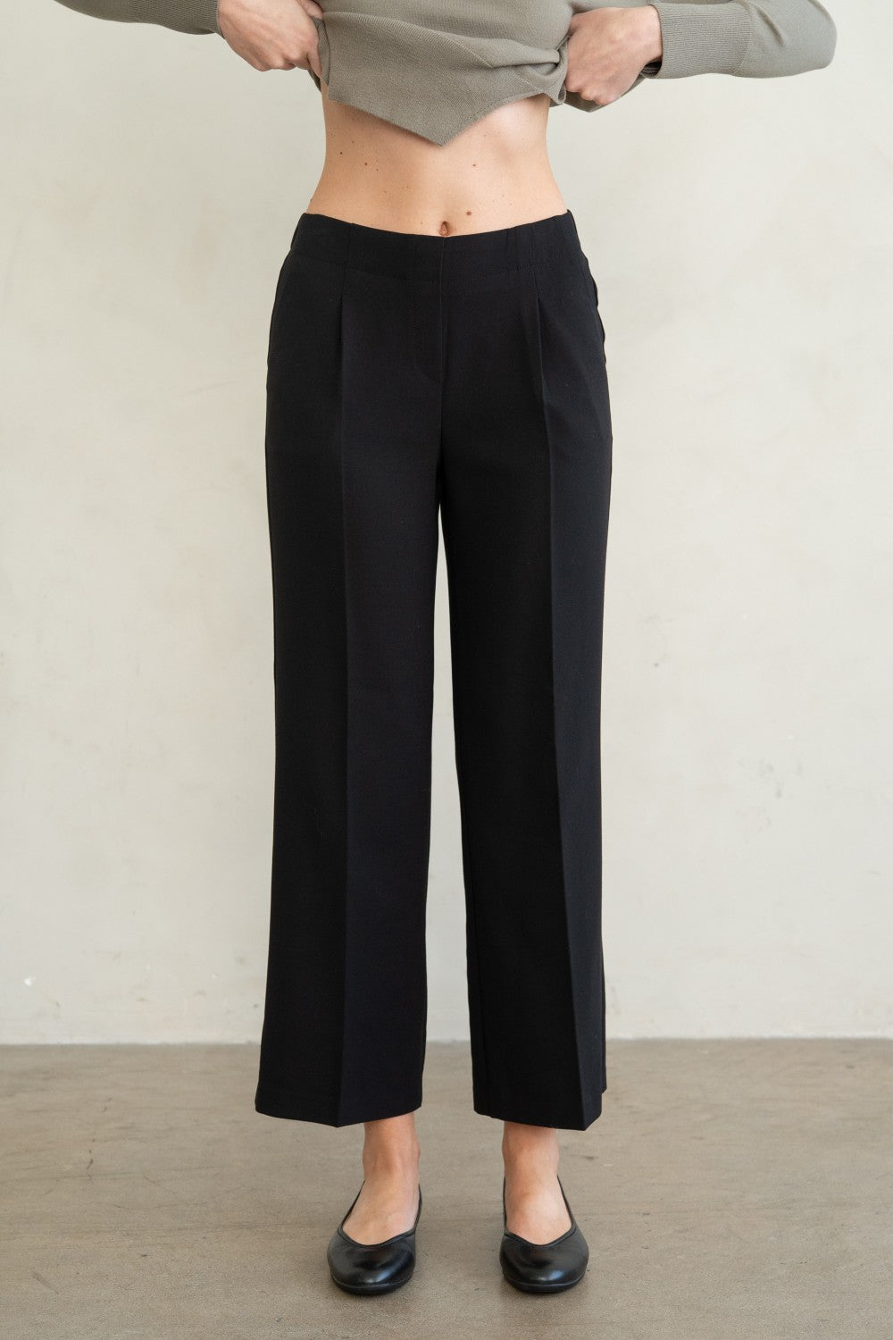 A model wearing black wide-leg cropped trousers, paired with a gray cropped top and black flats, standing against a neutral off-white wall.