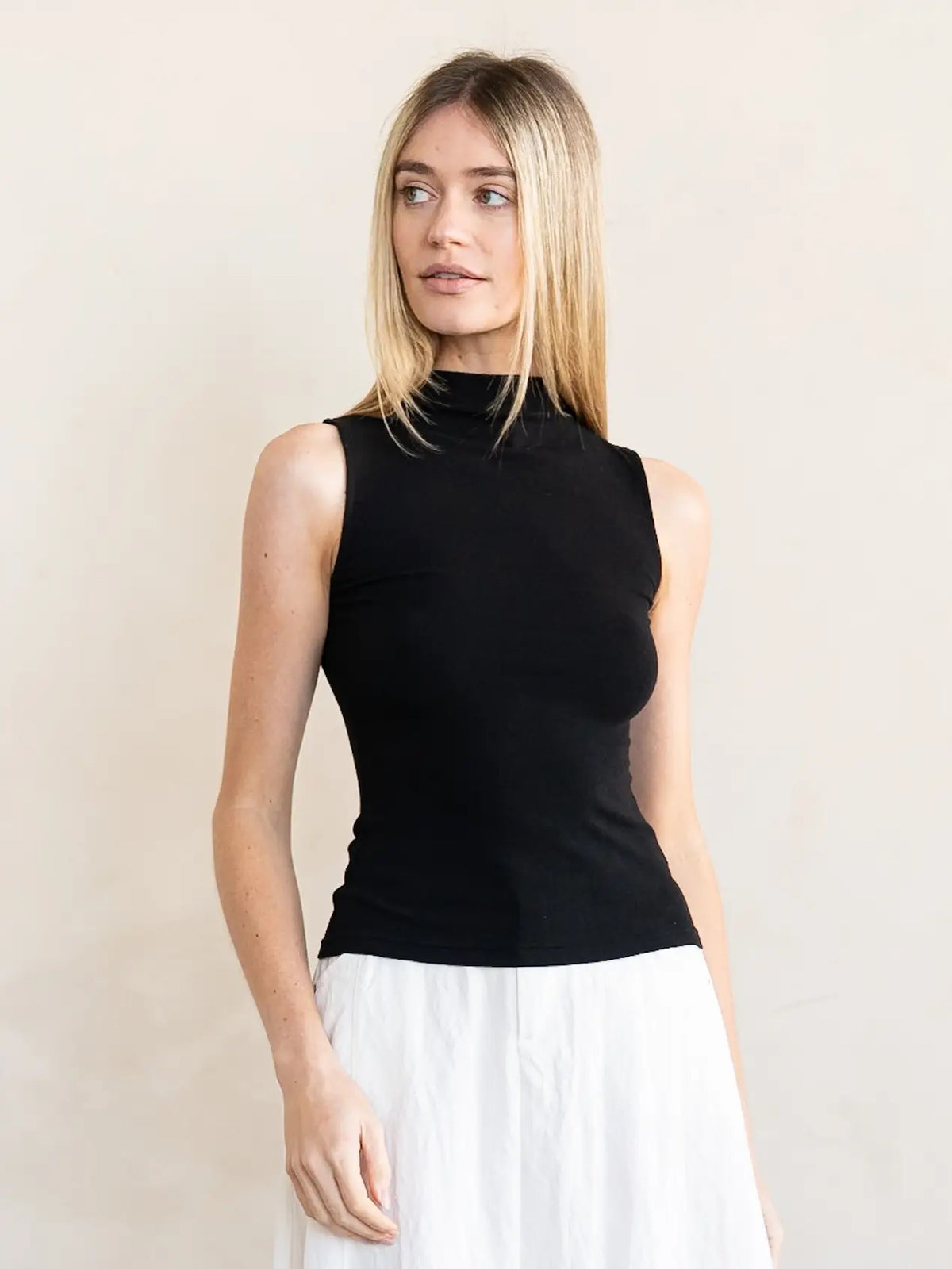 A woman modeling a sleeveless black mock neck top paired with a white skirt, standing against a plain beige wall.