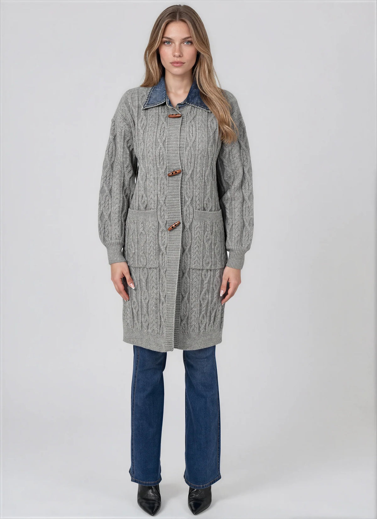 A woman modeling a gray cable-knit cardigan with a blue denim collar, brown toggle closures, and front patch pockets, paired with blue jeans and black shoes, against a light gray studio background.