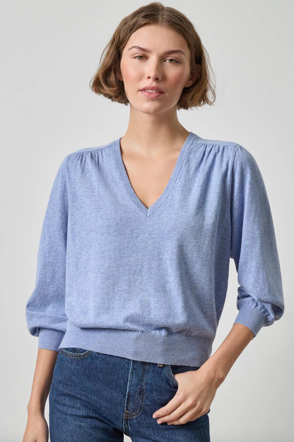 A woman wearing a light blue heathered knit sweater with a V-neck and gathered sleeves, paired with dark blue jeans, against a plain light gray studio background.