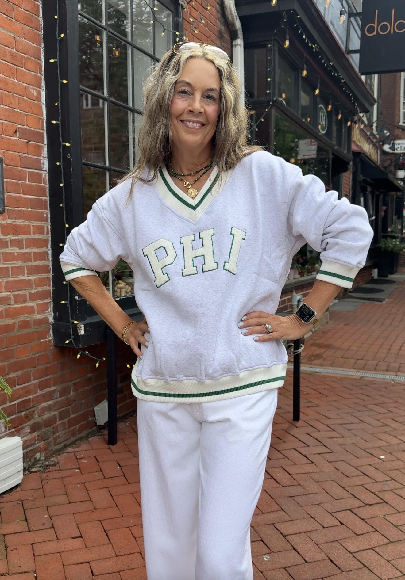 A woman stands outdoors on a brick sidewalk wearing a white V-neck sweatshirt with green trim and the letters 'PHI' across the chest, paired with white sweatpants.
