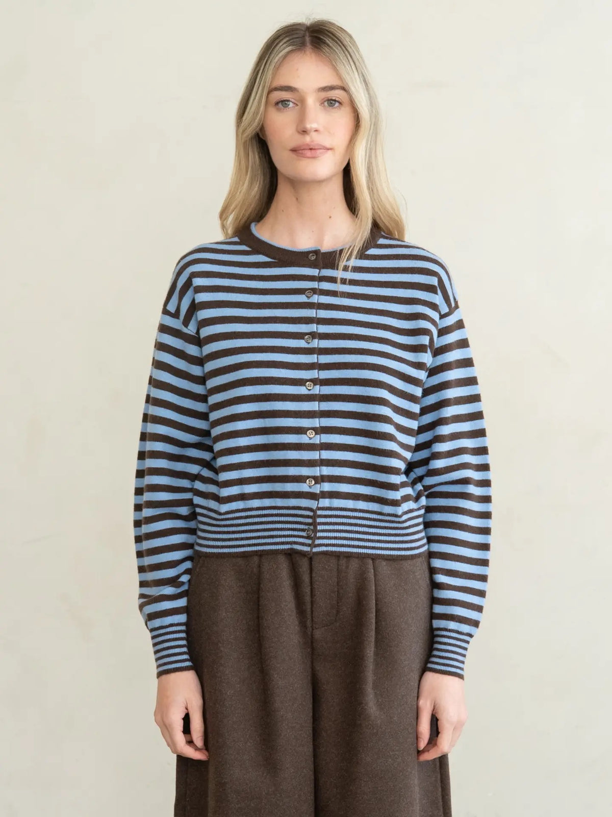 A woman modeling a cropped, long-sleeve knit cardigan with horizontal dark brown and light blue stripes, featuring a dark brown crew neck and button-front closure, paired with brown wide-leg trousers against a neutral cream background.