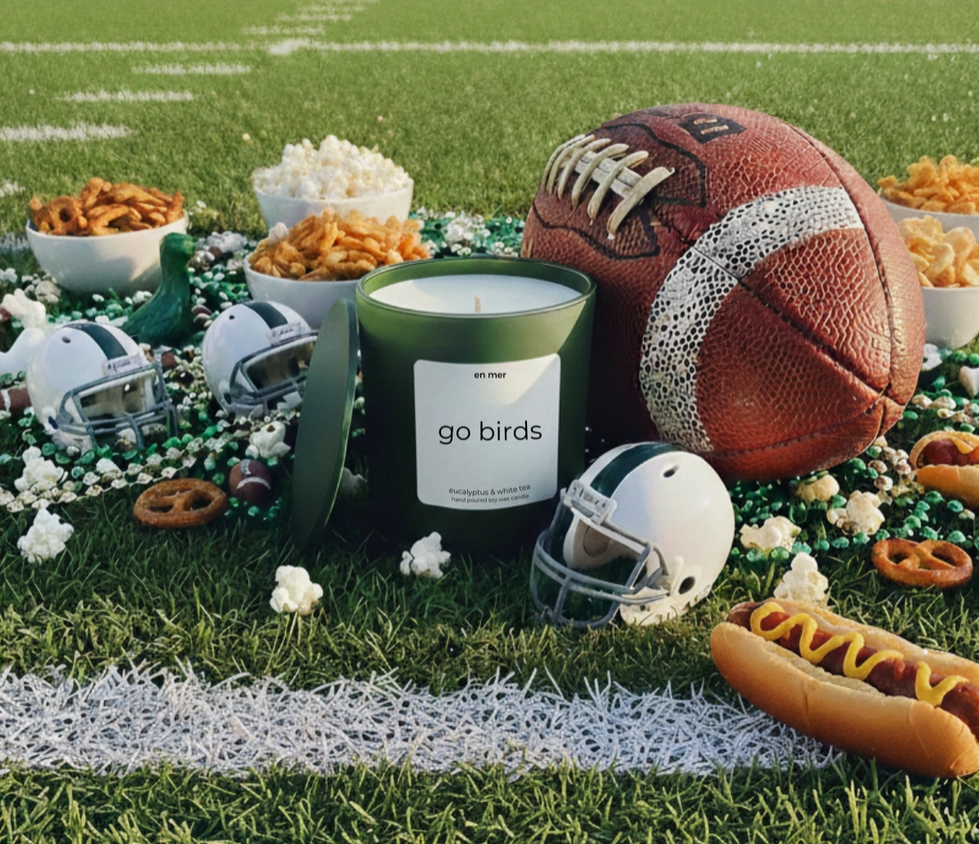 A green candle jar with a white label reading 'go birds' is styled on a football field with a brown football, white helmets, popcorn, pretzels, beans, mustard, and hot dogs.