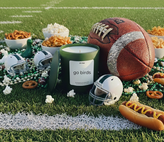 A green candle jar with a white label reading 'go birds' is styled on a football field with a brown football, white helmets, popcorn, pretzels, beans, mustard, and hot dogs.
