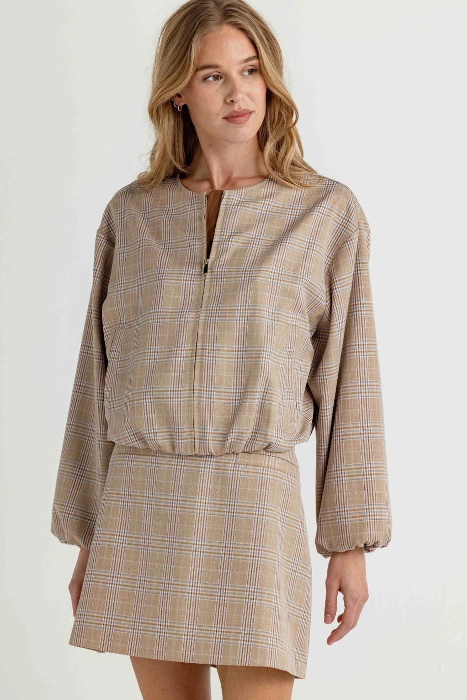 A woman wearing a beige plaid mini dress with a half-zip neckline and long balloon sleeves, photographed against a plain light background.