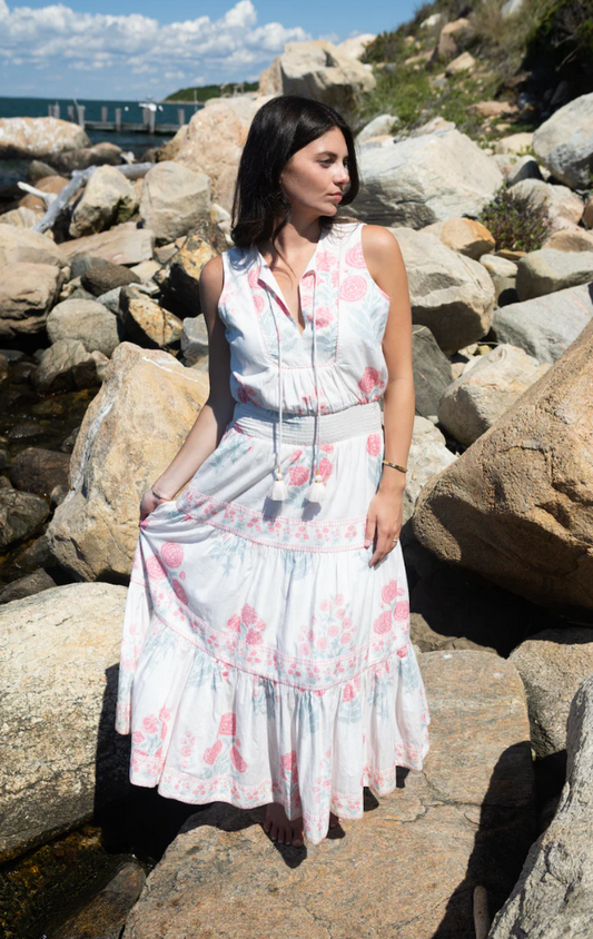 A woman wearing a sleeveless white maxi dress with a pink and light blue floral print stands on large rocks outdoors near the water, holding out one side of the dress to show its full tiered length.