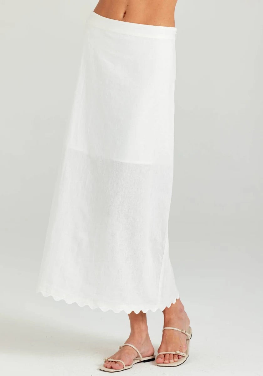 A white maxi skirt with a scalloped hem, shown on a model wearing silver flat sandals against a light gray studio background.