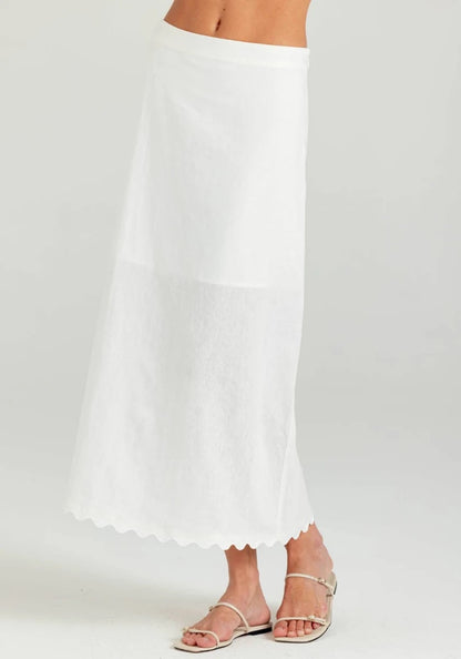 A white maxi skirt with a scalloped hem, shown on a model wearing silver flat sandals against a light gray studio background.