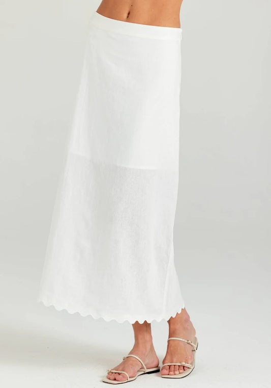 A white maxi skirt with a scalloped hem, shown on a model wearing silver flat sandals against a light gray studio background.