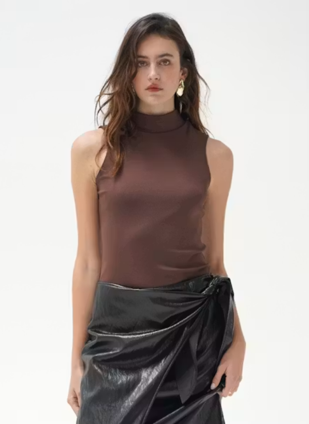 A woman modeling a sleeveless mock-neck top in dark brown paired with a shiny black draped wrap skirt, set against a plain white studio background.