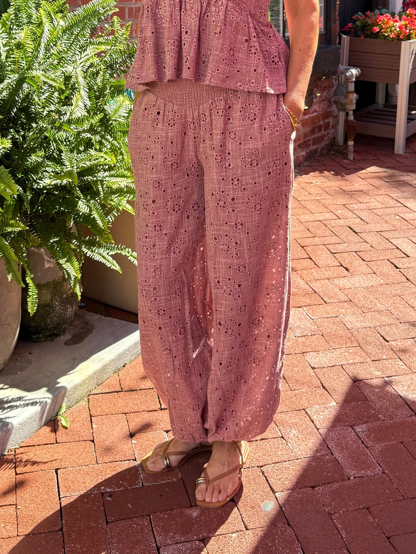 A woman wearing a dusty rose pink two-piece matching set consisting of a sleeveless peplum top and wide-leg trousers, standing on a red brick sidewalk in front of a brick building.