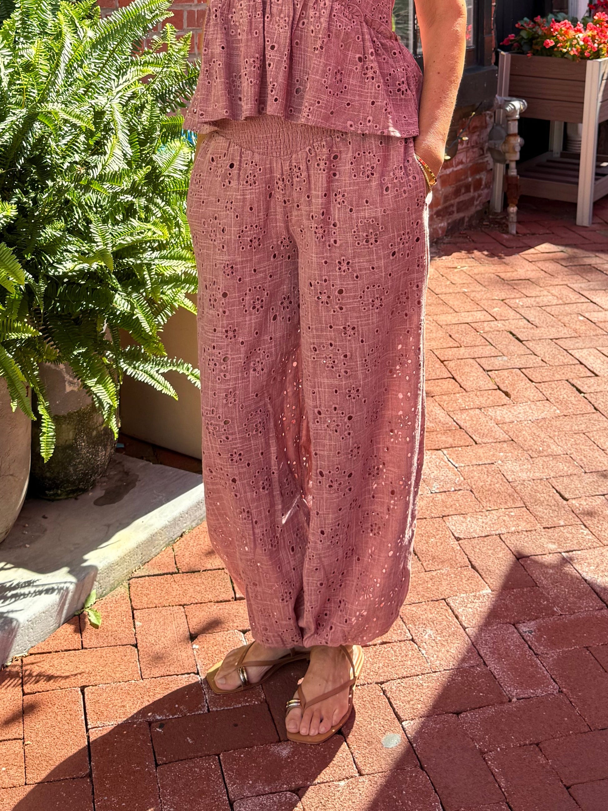 A woman wearing a dusty rose pink two-piece matching set consisting of a sleeveless peplum top and wide-leg trousers, standing on a red brick sidewalk in front of a brick building.