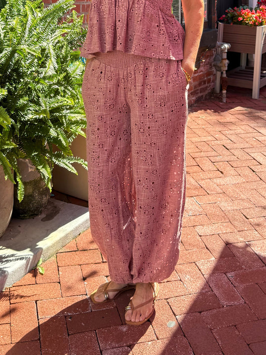 A woman wearing a dusty rose pink two-piece matching set consisting of a sleeveless peplum top and wide-leg trousers, standing on a red brick sidewalk in front of a brick building.