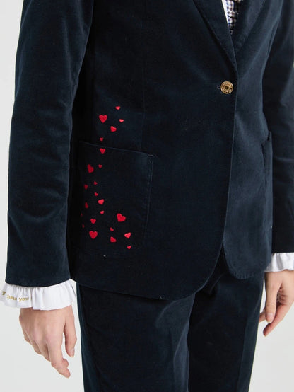 A close-up of a dark navy velvet blazer with red embroidered hearts on the pocket, worn by a model with a white ruffled shirt cuff visible.