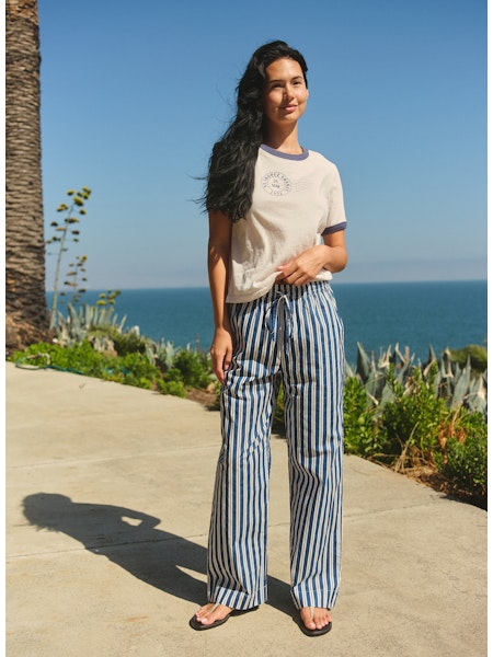 Woman standing on a path by the ocean with palm trees and clear blue sky.