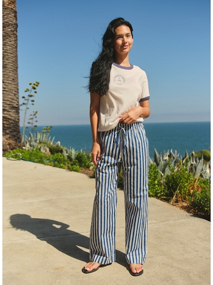 Woman standing on a path by the ocean with palm trees and clear blue sky.