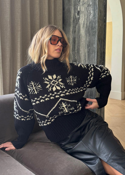 Woman wearing a dark sweater with white patterns, sitting on a couch indoors.