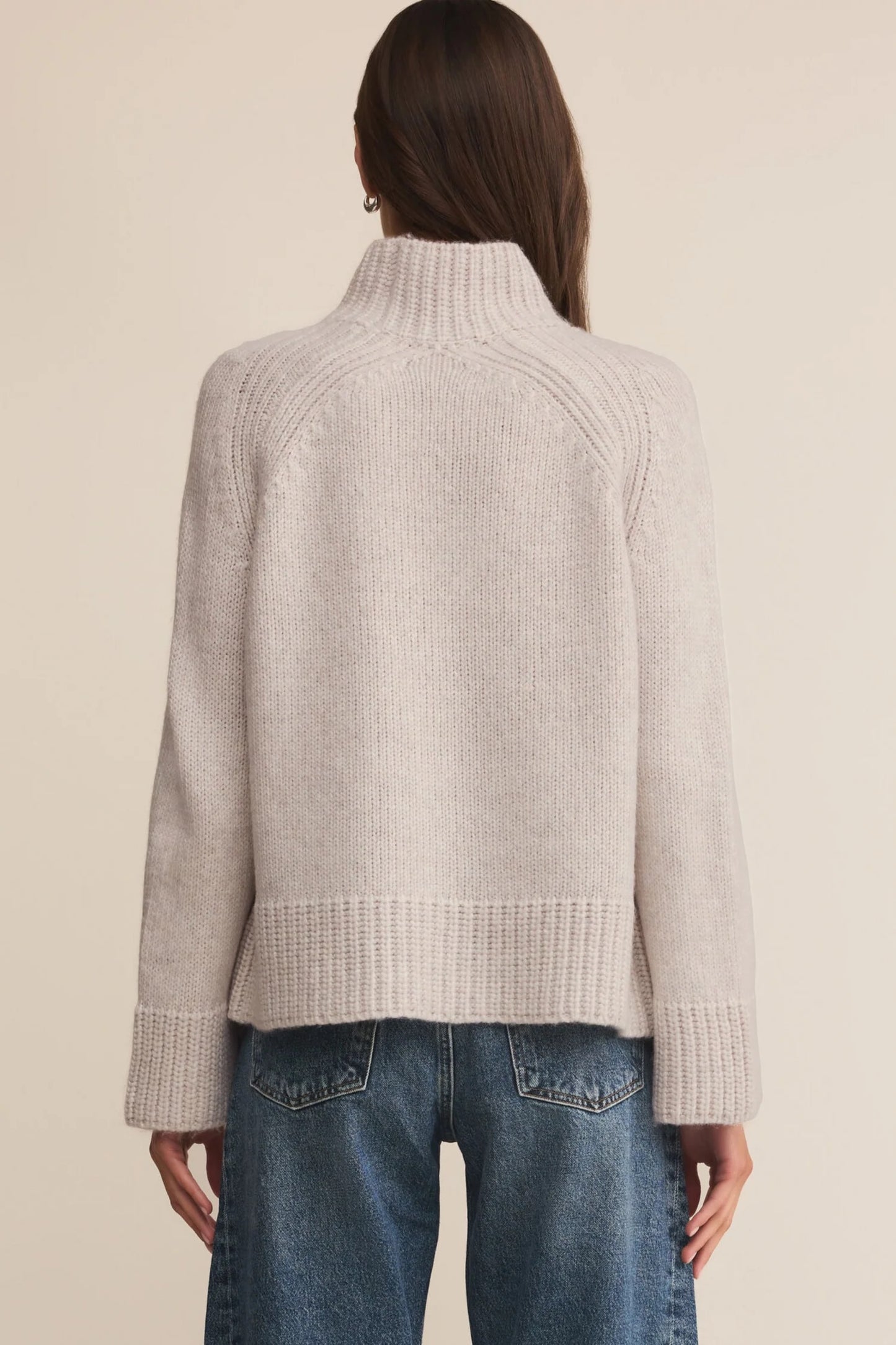 Person wearing a light gray ribbed sweater and blue jeans on a beige background