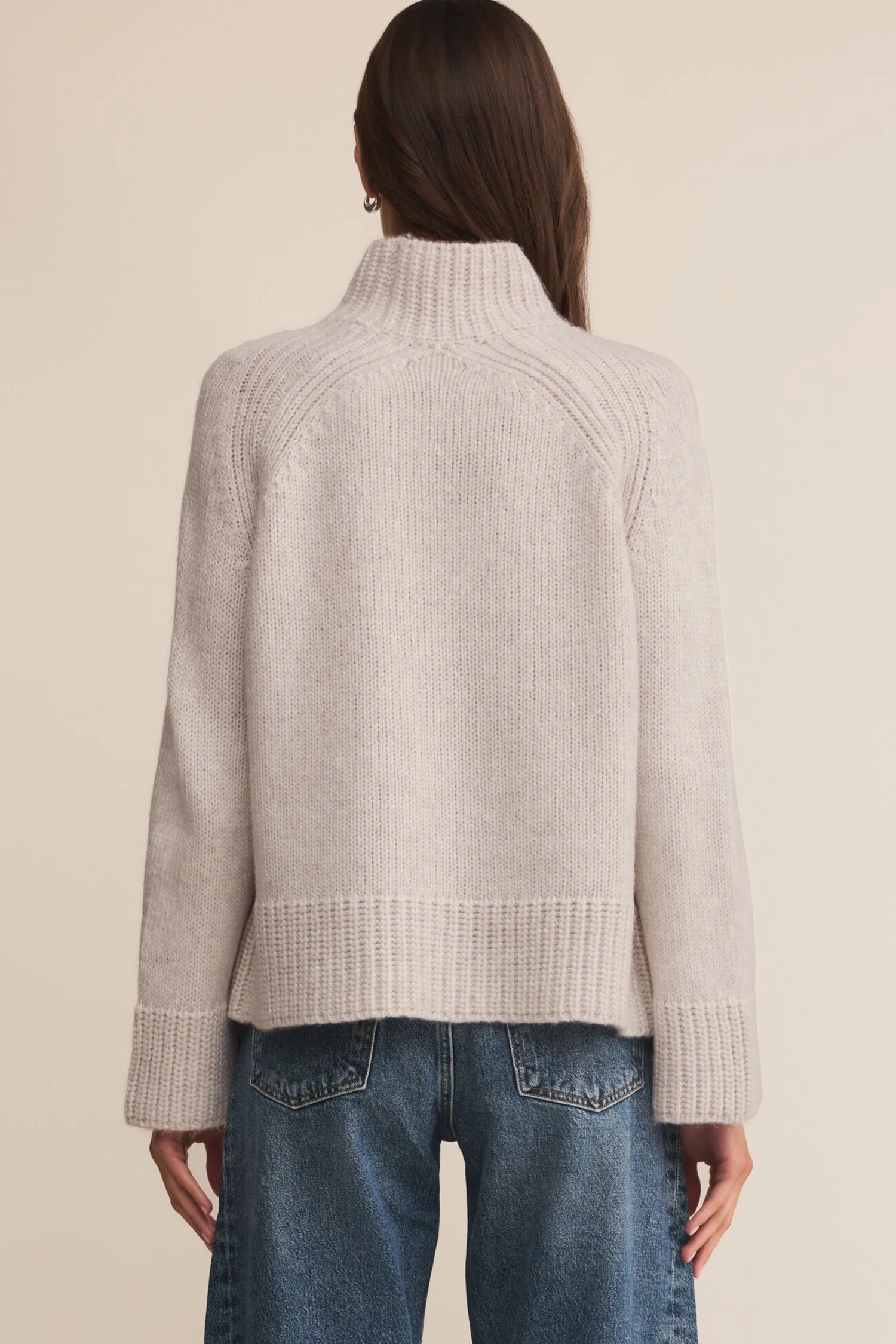 Person wearing a light gray ribbed sweater and blue jeans on a beige background