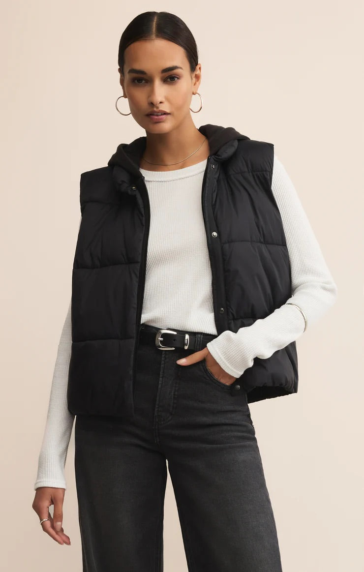 Woman wearing a black puffer vest over a white long-sleeve shirt on a beige background