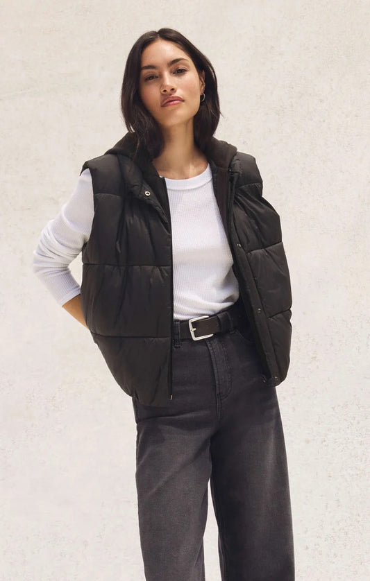 Woman wearing a black puffer vest over a white shirt and gray pants on a light background