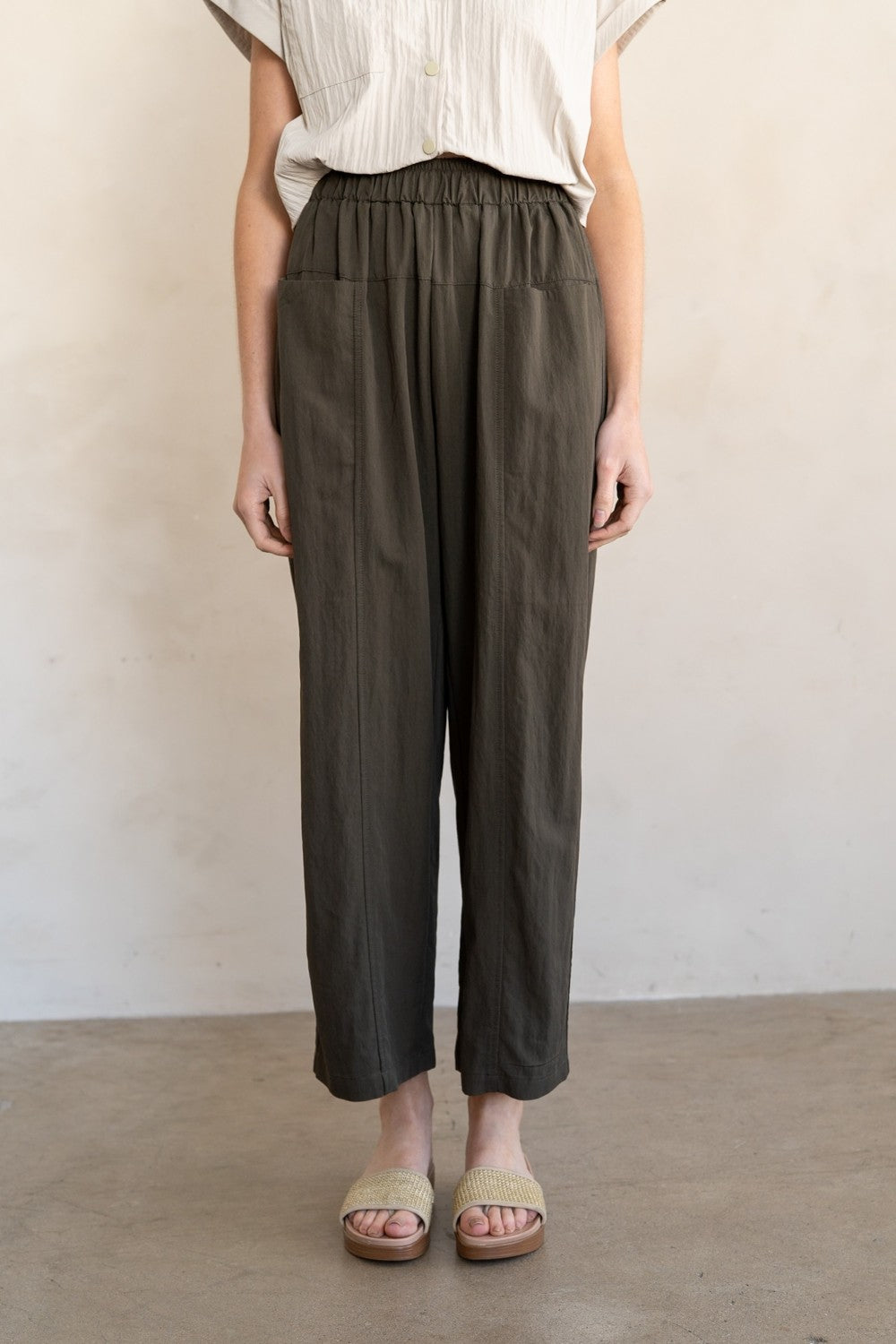 A person wearing loose, high-waisted olive green cropped trousers with an elastic waistband, paired with a cream pleated top and beige slide sandals, standing against a plain light beige wall and concrete floor.