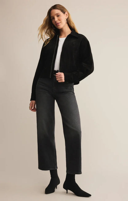 Woman wearing a black jacket, white shirt, gray pants, and black boots on a beige background