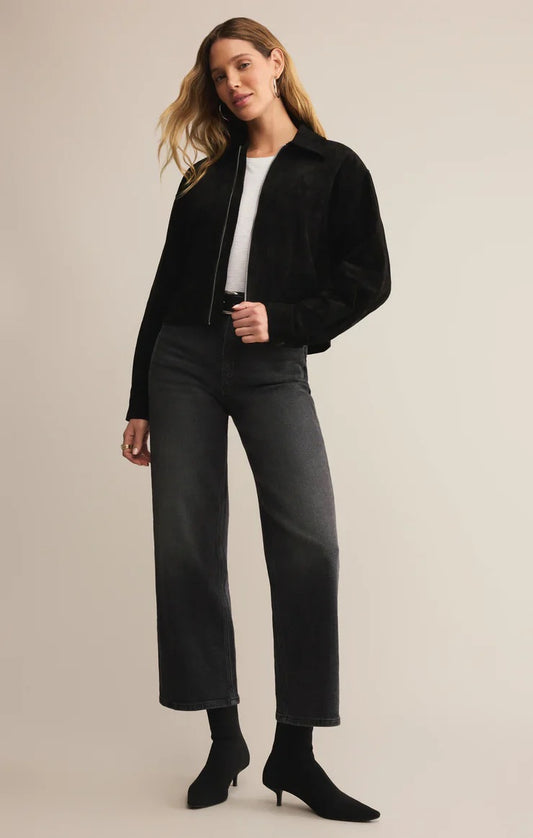 Woman wearing a black jacket, white shirt, gray pants, and black boots on a beige background
