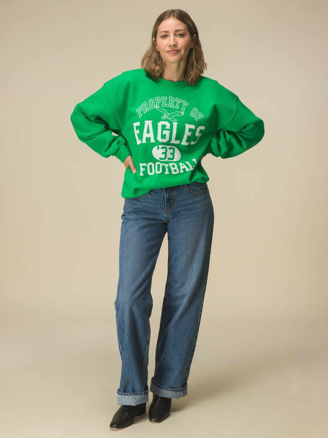 Women in front of a beige background in a green crewneck sweatshirt that says "property of eagles football"