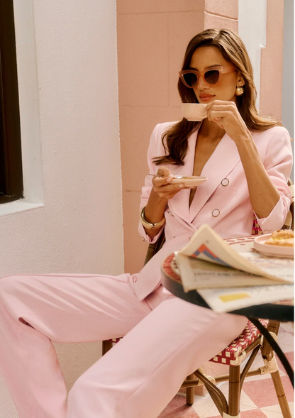 Woman in a pink suit sitting at an outdoor cafe, sipping coffee.