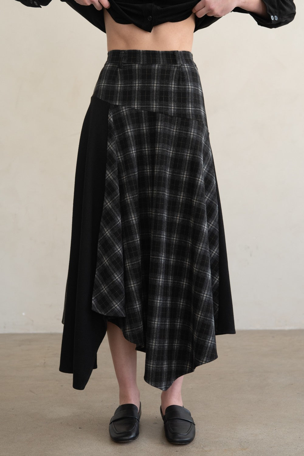 A person wearing a black and gray plaid asymmetrical skirt with solid black side panels, paired with black loafers, against a plain beige wall and floor.