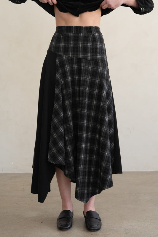 A person wearing a black and gray plaid asymmetrical skirt with solid black side panels, paired with black loafers, against a plain beige wall and floor.