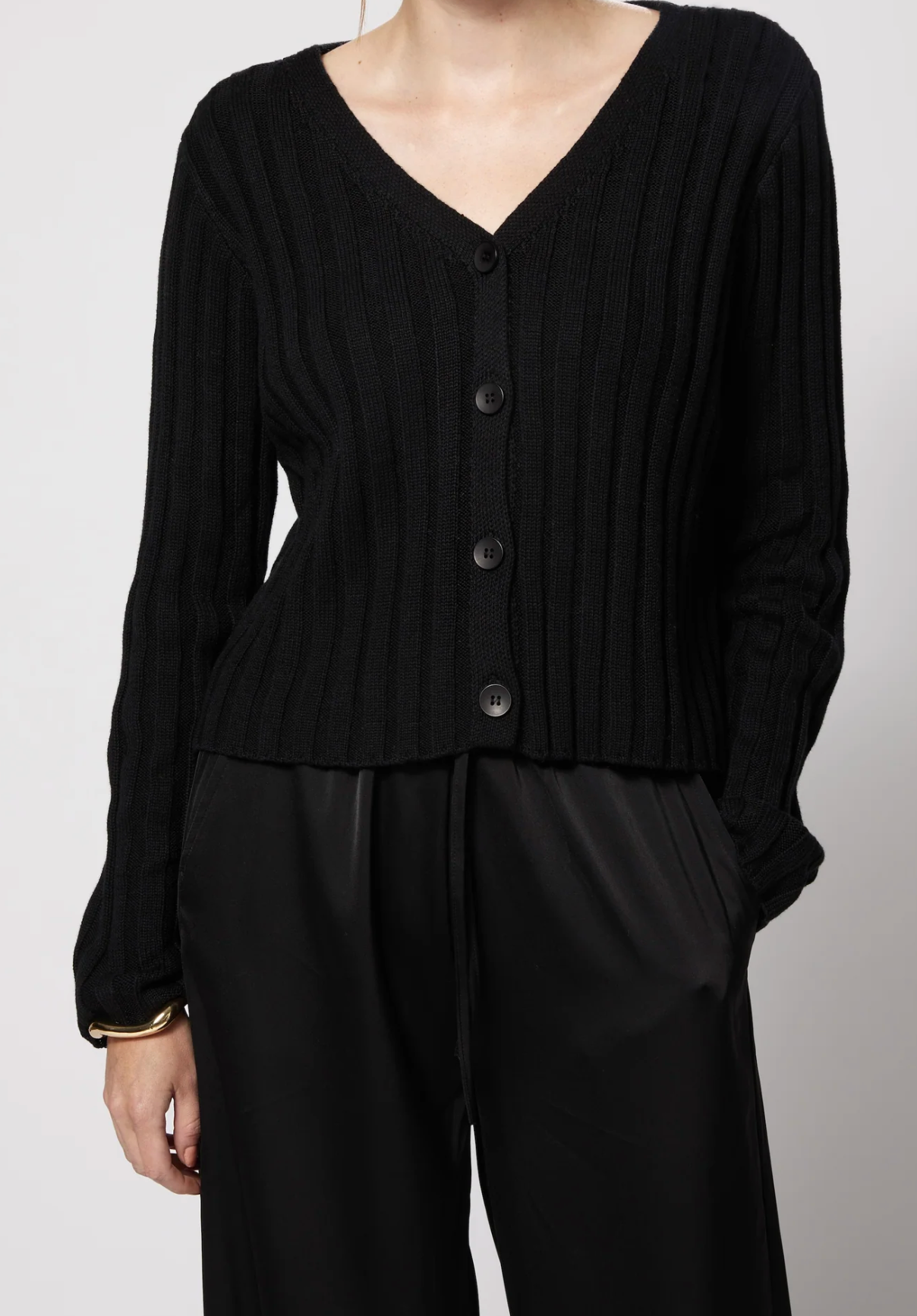 A person wearing a black ribbed knit cardigan with a V-neck and front buttons, paired with black pants, photographed against a light gray studio background.