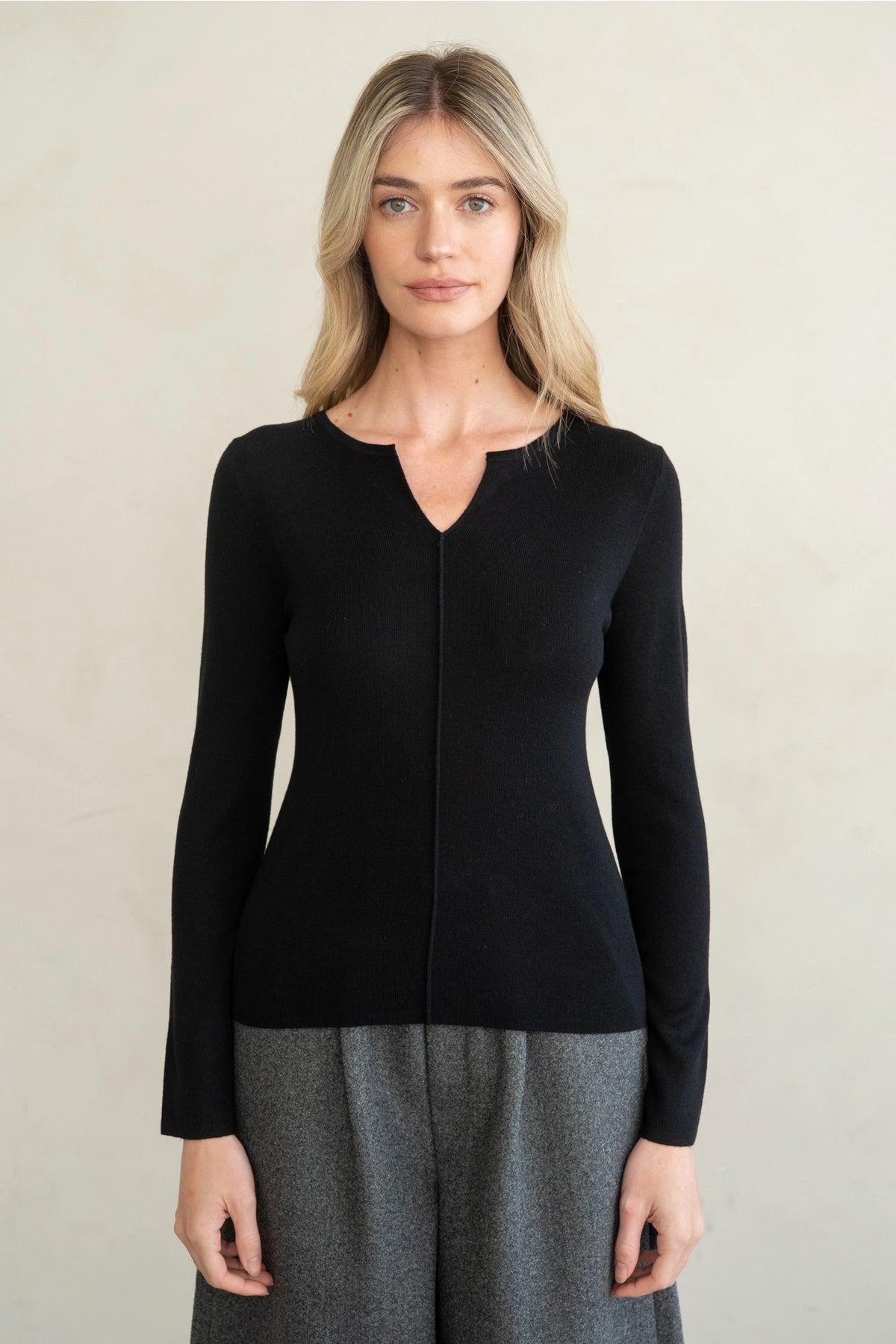 A woman modeling a black long-sleeve knit top with a split V-neckline and front zipper, paired with gray wide-leg trousers, against a plain light beige wall.