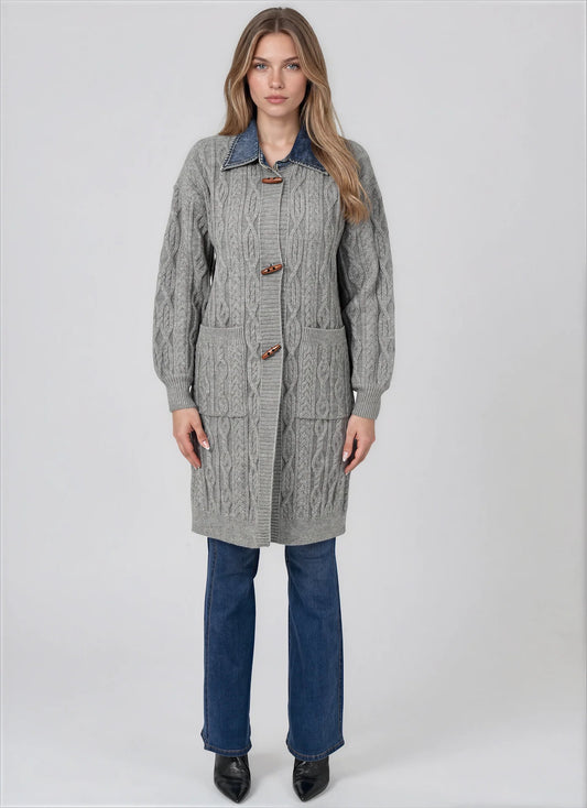 A woman modeling a gray cable-knit cardigan with a blue denim collar, brown toggle closures, and front patch pockets, paired with blue jeans and black shoes, against a light gray studio background.