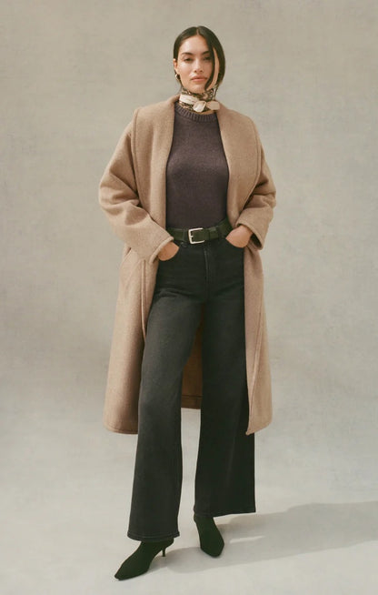 Woman wearing a beige coat, brown sweater, and black pants on a plain background