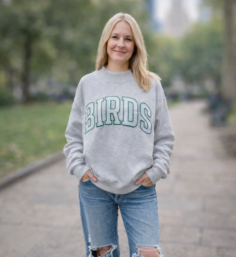 A woman stands on a blurred park path wearing a light gray crewneck sweatshirt with 'BIRDS' printed across the chest in green varsity-style lettering, paired with distressed blue jeans.