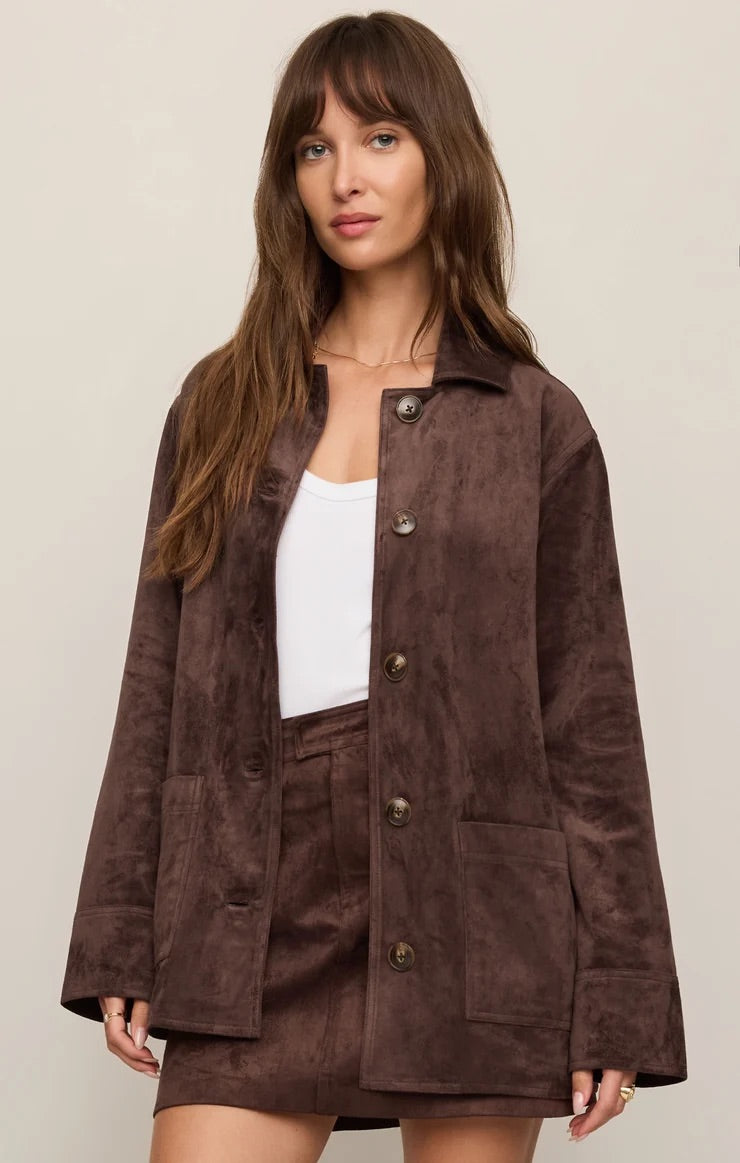 Woman wearing a brown suede jacket over a white top and brown shorts on a beige background