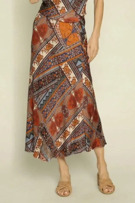 A woman modeling a sleeveless halter-neck maxi dress with a complex geometric and floral pattern in orange, brown, blue, and white. She is also wearing a wide-brimmed tan hat and tan sandals, standing against a plain light beige background.