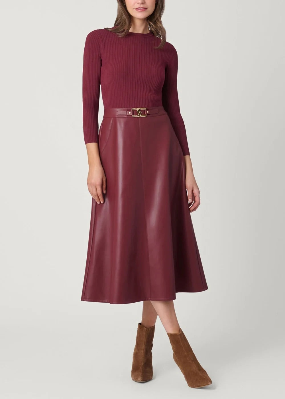 A woman models a burgundy ribbed knit long-sleeve top paired with a matching faux-leather midi skirt featuring a gold-tone belt, styled with brown suede ankle boots against a light gray studio background.