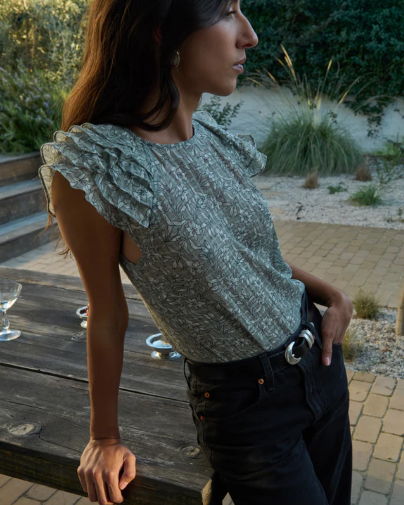 Woman wearing a patterned top and dark pants standing outdoors on a wooden deck.