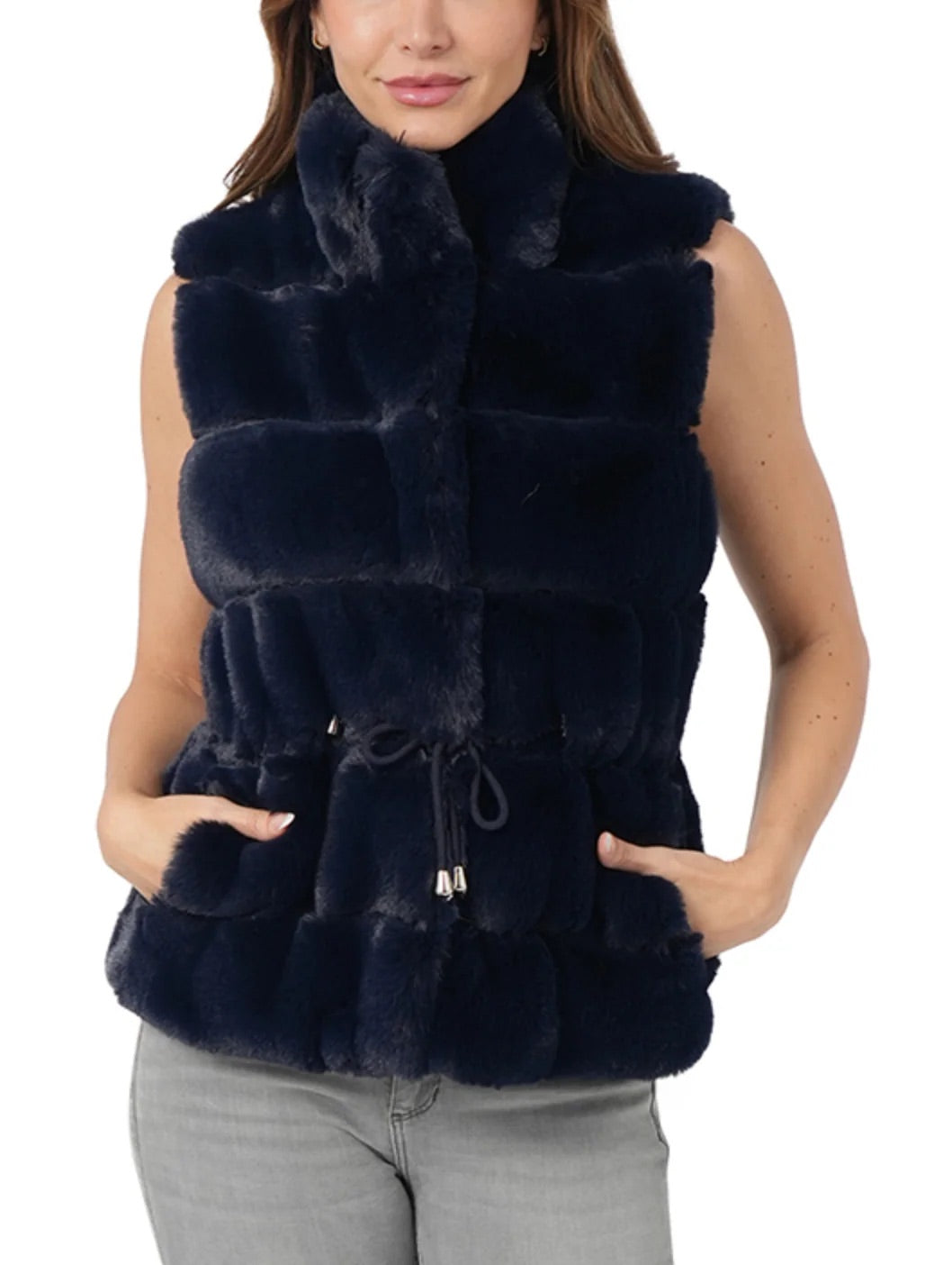 A woman wearing a sleeveless, dark navy faux fur vest with a high collar and drawstring waist, paired with gray jeans, against a plain white background.