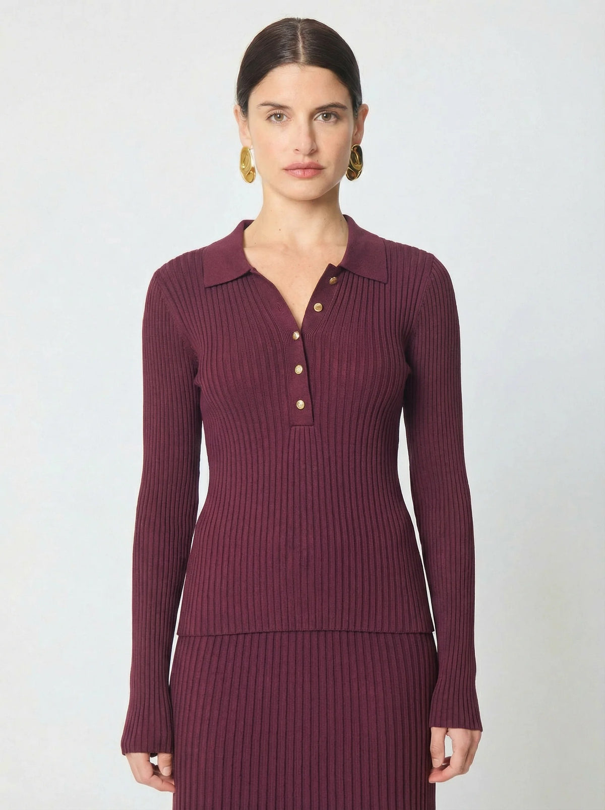 A woman modeling a ribbed long-sleeve polo shirt in deep burgundy with gold buttons, paired with a matching ribbed skirt, against a light gray studio background.
