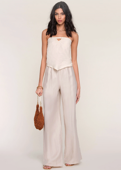 Woman wearing a strapless white top and beige wide-leg pants holding a woven bag on a plain background