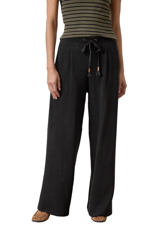 A model wearing black wide-leg pleated pants with a drawstring waist, paired with a striped top and brown flat shoes, shown against a plain white background.