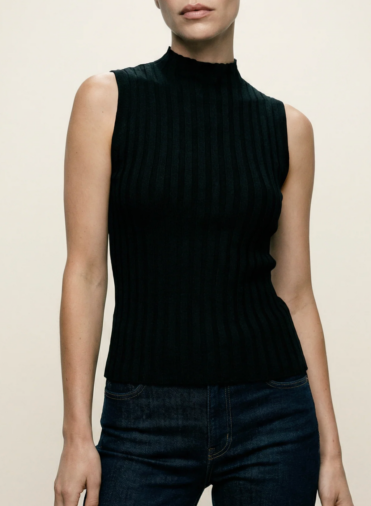 A model wearing a sleeveless, ribbed black mock neck top paired with dark blue jeans, shown from the lips down against a plain light beige background.