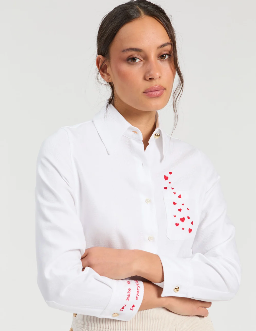 A woman wearing a white long-sleeve button-up shirt with red heart embroidery on the chest pocket and cuff, paired with a cream ribbed top, against a light gray studio background.